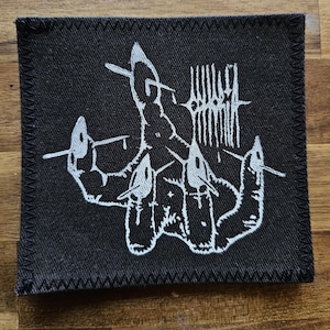 Peut inclure: Écusson carré noir avec un motif brodé blanc. Le motif représente une main avec de multiples lignes et formes. L'écusson a une bordure noire avec des coutures blanches. Le fond est une surface en bois.