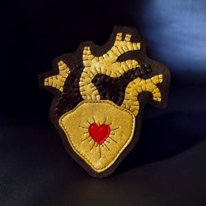 May include: A handcrafted patch featuring a stylized anatomical heart design. The patch has a dark brown background with gold-colored veins and a gold heart-shaped center. A small red heart is in the center of the gold heart.