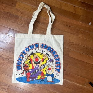 May include: A cream-colored canvas tote bag featuring a colorful graphic print with the text HUEYTOWN SPRINGFEST. The design includes three cartoon characters, a purple electric guitar, and musical notes on a blue oval base.