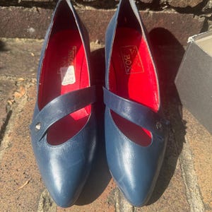 May include: A pair of navy blue leather pumps featuring a pointed toe, a single diagonal strap across the vamp with a small silver-tone stud, and a vibrant red interior lining. The insole displays the text P.S. BOX in a rectangular gold-tone label.