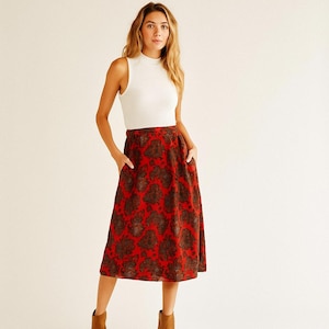 May include: A woman's outfit featuring a white sleeveless top and a red midi skirt. The skirt has a brown floral pattern and pockets. The outfit is suitable for casual or semi-formal occasions.