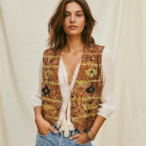 May include: A brown and gold patterned vest with black detailing. The vest is worn over a white blouse with a tassel tie. The model is wearing blue jeans and gold bracelets. The vest has a geometric design.