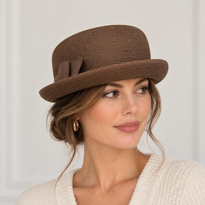 May include: A dark brown structured hat with a rounded crown and a short, slightly upturned brim. The hat features a textured, ribbed surface and a small, matching brown fabric bow detail positioned on the side of the crown above the brim.