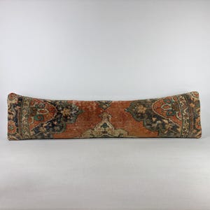 May include: A long, rectangular bolster pillow featuring a detailed, multi-colored woven pattern in shades of burnt orange, deep charcoal, and muted cream. The textile displays a symmetrical, intricate design with floral and geometric motifs.