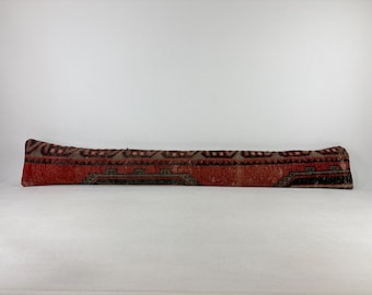Peut inclure: Un long coussin rectangulaire avec un motif. Le coussin présente un fond rouge avec des motifs géométriques noirs, marron et beige. Le coussin est probablement fait de tissu tissé et est un article décoratif.