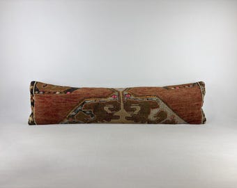 May include: A long, rectangular lumbar pillow featuring a woven textile cover in shades of terracotta, brown, and cream. The fabric displays a symmetrical, geometric pattern with small accents of pink and yellow throughout the design.