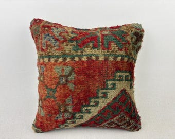 May include: A square throw pillow featuring a textured, woven surface with a multi-colored pattern. The design incorporates deep red, muted green, and beige tones in a geometric arrangement, showcasing a dense, pile-like fabric construction.