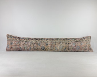 May include: A long, rectangular lumbar pillow featuring a faded, vintage-style textile cover. The fabric displays a muted color palette of dusty pink, beige, and deep blue, decorated with an intricate, repeating floral and vine pattern.