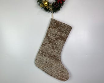Puede incluir: Un calcetín navideño con un patrón tejido marrón y beige cuelga de una corona decorativa. La corona presenta adornos dorados, agujas de pino verdes y bayas rojas. El calcetín está hecho de una tela texturizada.