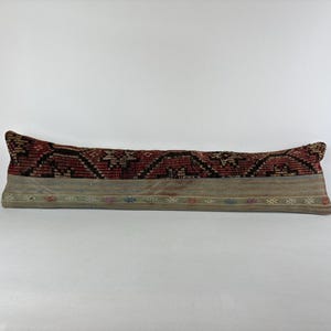 May include: A long, rectangular pillow with a patterned design. The top section features a red and black geometric pattern, while the bottom has a tan base with a decorative border. The pillow is designed for decorative use.