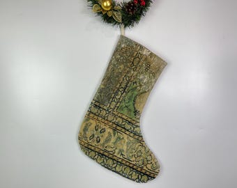 Puede incluir: Un calcetín navideño con un diseño de alfombra vintage. El calcetín presenta un patrón beige, verde y negro con elementos florales y geométricos. Una pequeña corona navideña con detalles dorados y rojos cuelga encima.