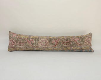 May include: A long, rectangular pillow with a colorful, patterned fabric. The fabric has a faded, vintage look with shades of pink, blue, green, and beige.