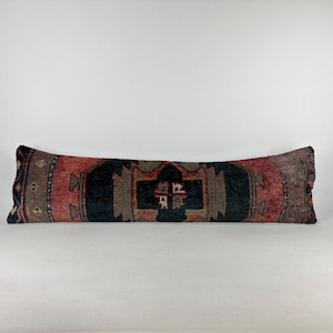 May include: A long, rectangular pillow with a patterned design. The pillow features a central black section with geometric shapes, flanked by red and brown sections. The edges have a darker, faded appearance, suggesting an aged or vintage aesthetic.