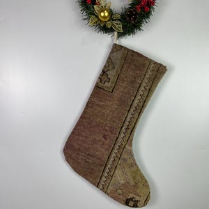 May include: A Christmas stocking made from a patterned rug, in shades of brown, beige, and green. The stocking is hanging from a Christmas wreath with red berries, pinecones, and a gold ornament.