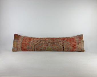 May include: A long, rectangular lumbar pillow featuring a woven textile cover with a distressed, faded appearance. The fabric displays a complex pattern in shades of burnt orange, muted red, beige, and hints of green, with dark black outlines.