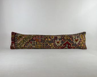 May include: A long, rectangular pillow with a patterned design. The pillow features a dark gray background with red, yellow, and green floral and geometric patterns. The ends have a red border with intricate designs.