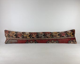 Puede incluir: Un cojín lumbar largo y rectangular con un patrón textil tejido en tonos rojo, negro, beige y amarillo. El diseño consiste en motivos geométricos repetitivos y formas florales estilizadas dispuestas en una banda horizontal sobre la superficie.
