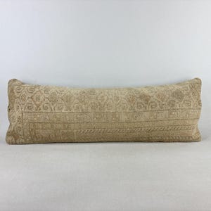 May include: A long, rectangular lumbar pillow featuring a muted beige and tan color palette. The fabric displays a detailed, woven geometric pattern with intricate borders and repeating diamond motifs across the entire surface of the cushion.