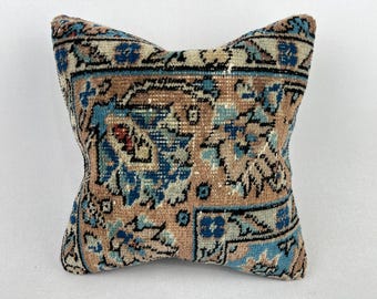 May include: Square decorative pillow with a vintage rug design. The pillow features a beige background with blue and brown floral patterns. The edges are bordered with a dark blue and black design.