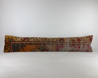 Puede incluir: Un cojín lumbar largo y rectangular con un patrón textil de estilo vintage descolorido en tonos naranja quemado, rojo intenso y gris carbón. La tela muestra intrincados motivos geométricos y florales con una textura desgastada.