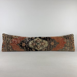 May include: A long, rectangular lumbar pillow featuring a faded, intricate pattern in shades of terracotta, black, and beige. The textile displays a central medallion design with ornate borders, showcasing a weathered, aged appearance throughout.