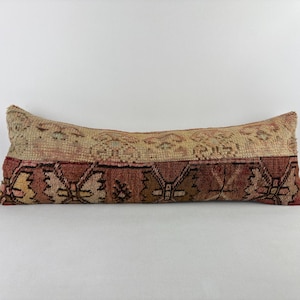 May include: A long, rectangular cushion with a patterned design. The top half is a light beige colour with a repeating pattern, while the bottom half is a darker red with a geometric design. The cushion is likely made of a woven fabric.
