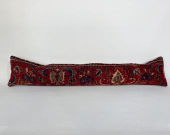 May include: A long, rectangular, red rug pillow with an intricate floral pattern in blue, green, and beige. The pillow is a decorative item, likely for a sofa or bed, and adds a touch of vintage charm.