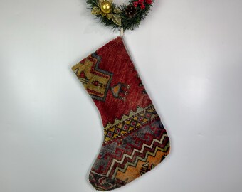 Puede incluir: Un calcetín navideño con un diseño estampado en rojo, dorado y gris. El calcetín cuelga de una corona decorativa con detalles dorados, agujas de pino y bayas rojas. El calcetín tiene un aspecto vintage, similar a una alfombra.
