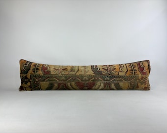 May include: A long, rectangular pillow with a vintage, patterned design. The pillow features a central panel with a repeating geometric pattern in shades of green, red, and beige. The ends have a darker, more intricate design. The pillow is approximately 36 inches long.