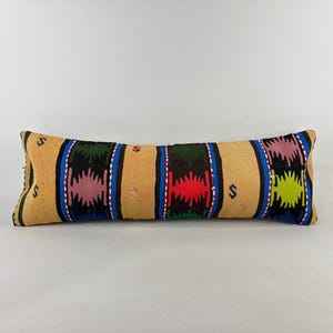 May include: A long rectangular lumbar pillow featuring a woven pattern with alternating tan, black, and blue vertical stripes. The black sections contain stylized geometric motifs in shades of pink, red, and lime green, accented by small white details.