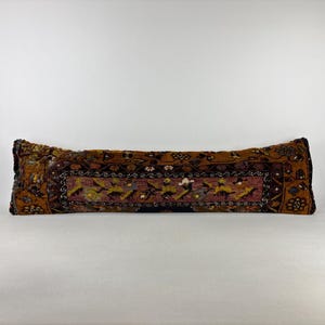 May include: A long, rectangular bolster pillow featuring a dark brown and rust-colored woven textile with a central panel displaying intricate floral and geometric patterns in shades of yellow, blue, and white, finished with dark brown fabric edges.