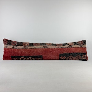 May include: A long, rectangular cushion with a patchwork design. The cushion features a mix of red, black, and tan colours with geometric and floral patterns. The cushion is designed for decorative use.