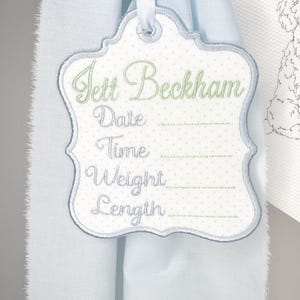 May include: Embroidered baby announcement tag with the name "Jett Beckham" in green script. The tag is white with a light blue border and polka dot pattern. It includes spaces for date, time, weight, and length.