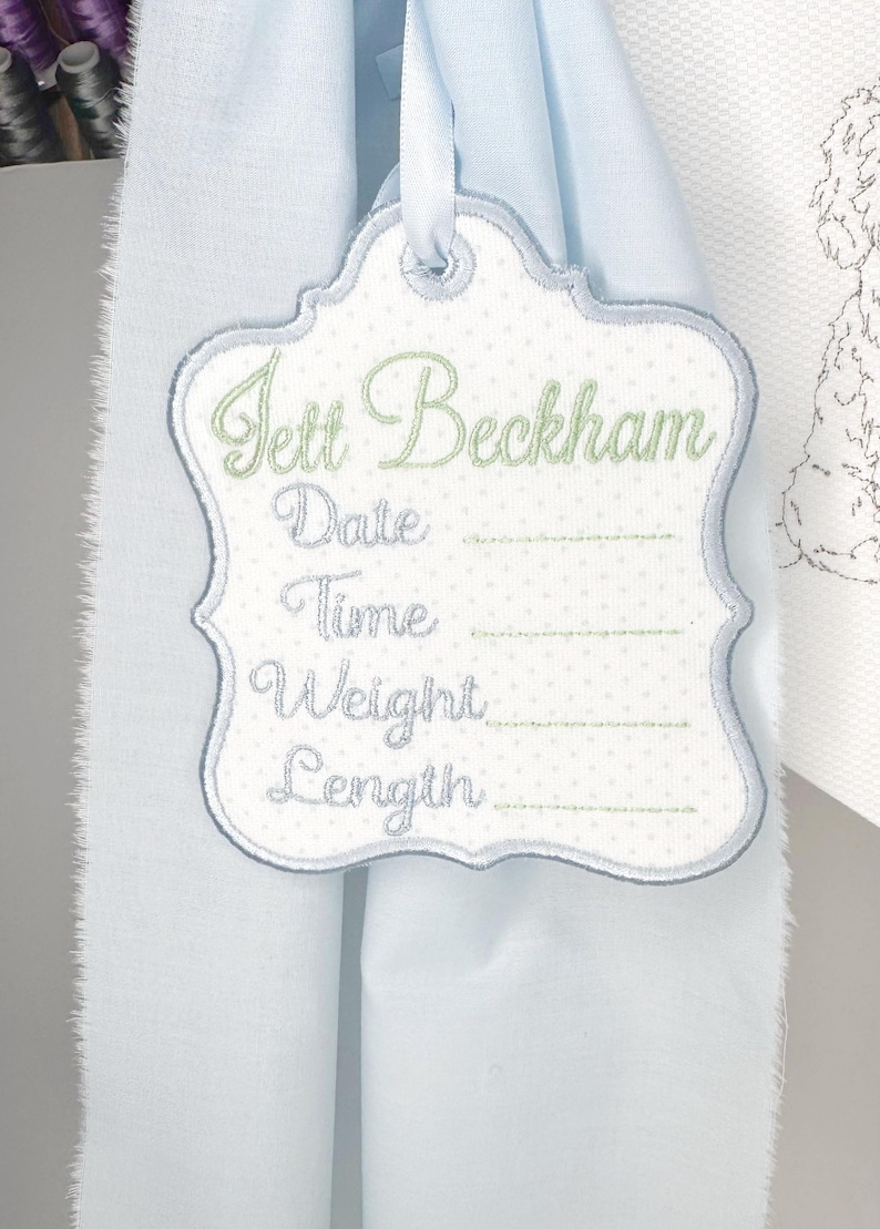 May include: Embroidered baby announcement tag with the name "Jett Beckham" in green script. The tag is white with a light blue border and polka dot pattern. It includes spaces for date, time, weight, and length.