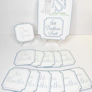 May include: Embroidered baby milestone signs with a light blue border. The signs include a large sign with a dog illustration, initials, and a name, plus smaller signs with months and year text.