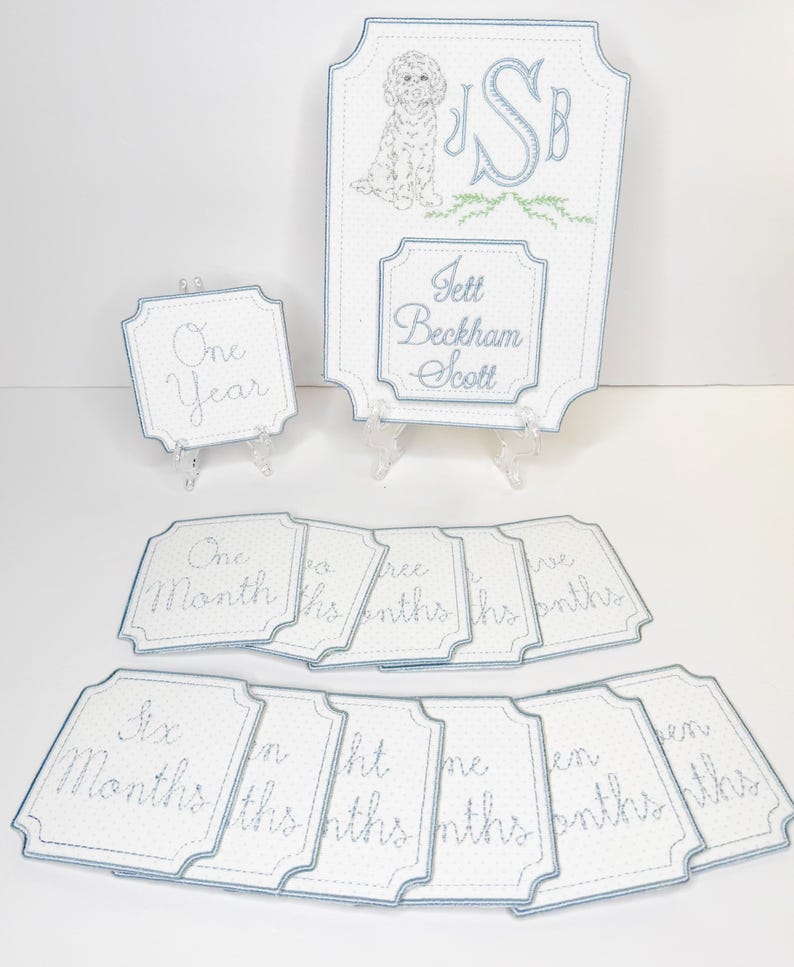 May include: Embroidered baby milestone signs with a light blue border. The signs include a large sign with a dog illustration, initials, and a name, plus smaller signs with months and year text.