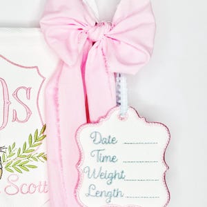 May include: A pink bow with a white ribbon is tied to a white tag with the words "Date", "Time", "Weight", and "Length" embroidered in blue. The tag is attached to a pink fabric.
