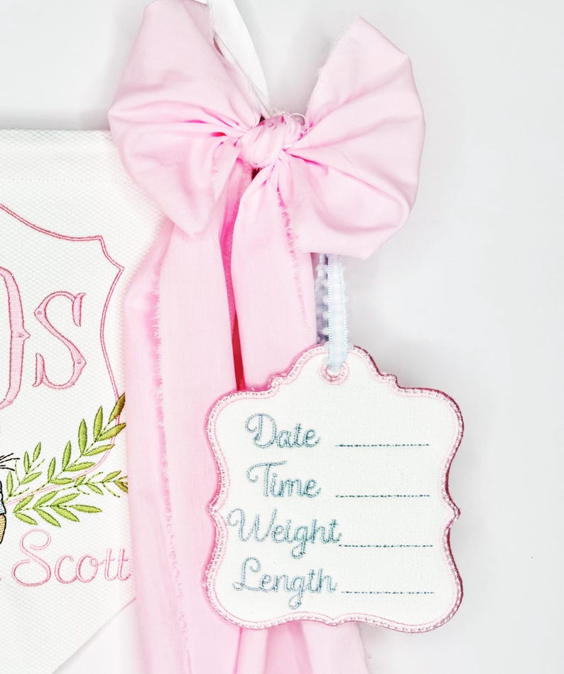 May include: A pink bow with a white ribbon is tied to a white tag with the words "Date", "Time", "Weight", and "Length" embroidered in blue. The tag is attached to a pink fabric.
