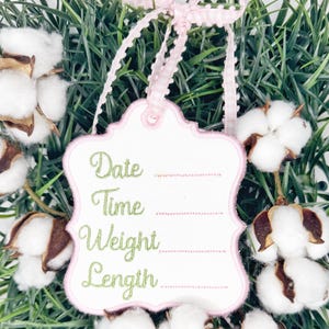 May include: A white tag with a pink scalloped border and a pink and white gingham ribbon. The tag has the words "Date", "Time", "Weight", and "Length" embroidered in green script. The tag is surrounded by cotton bolls and greenery.