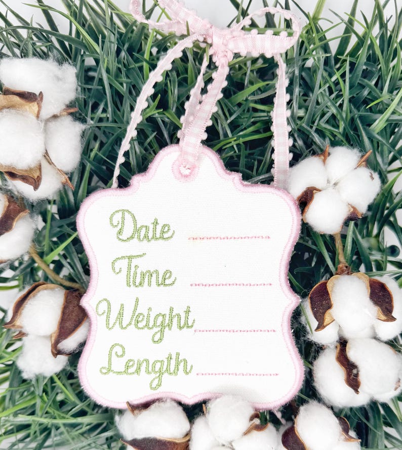 May include: A white tag with a pink scalloped border and a pink and white gingham ribbon. The tag has the words "Date", "Time", "Weight", and "Length" embroidered in green script. The tag is surrounded by cotton bolls and greenery.