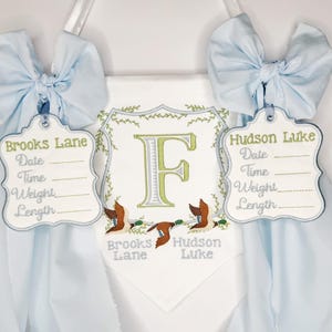 May include: A personalized baby banner with light blue bows and tags. The banner features embroidered details, including the names "Brooks Lane" and "Hudson Luke", along with spaces for date, time, weight, and length. A large letter "F" is centered.