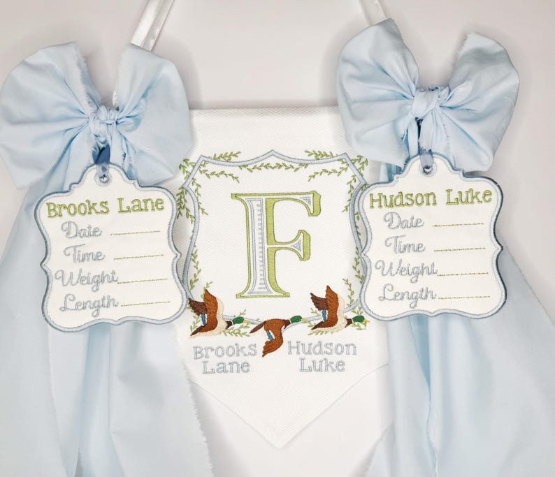 May include: A personalized baby banner with light blue bows and tags. The banner features embroidered details, including the names "Brooks Lane" and "Hudson Luke", along with spaces for date, time, weight, and length. A large letter "F" is centered.