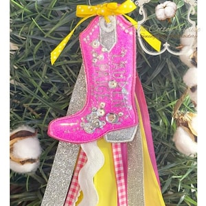 May include: A pink glitter cowboy boot decoration with silver detailing and decorative embellishments. The boot is decorated with rhinestones and a yellow bow. Ribbons in yellow, pink, and silver hang below the boot.