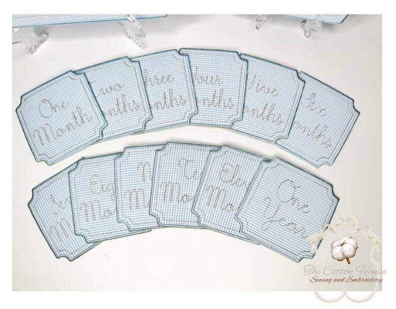 May include: A set of baby milestone cards with a blue and white gingham pattern. Each card is embroidered with a month or year, from "One Month" to "One Year". The cards have a decorative border.