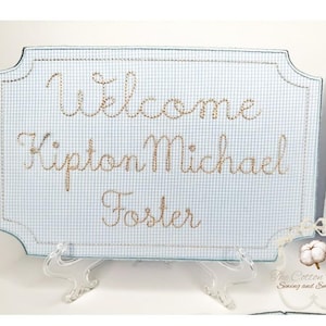 May include: A light blue and white gingham welcome sign embroidered with the name "Kipton Michael Foster" in a cursive font. The sign has a decorative border and is displayed on a clear stand.