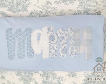 May include: A light blue pillow with the name "MOMMY" appliqued in various blue and white patterned fabrics. The letters are stitched onto the pillow, which is displayed on a patterned background.