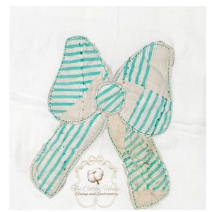 Może przedstawiać: Ręcznie robiona kokarda z tkaniny na białym tle. Kokarda ma naprzemienne paski w kolorze białym i turkusowym. Krawędzie są obrysowane przerywanym ściegiem. Tekst "The Cotton House Sewing and Embroidery" znajduje się na dole.