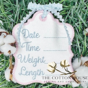 May include: White embroidered tag with pink trim and light blue text reading "Date, Time, Weight, Length." A light blue ribbon and cotton sprigs accent the tag. The text "THE COTTON HOUSE SEWING AND EMBROIDERY" is at the bottom.
