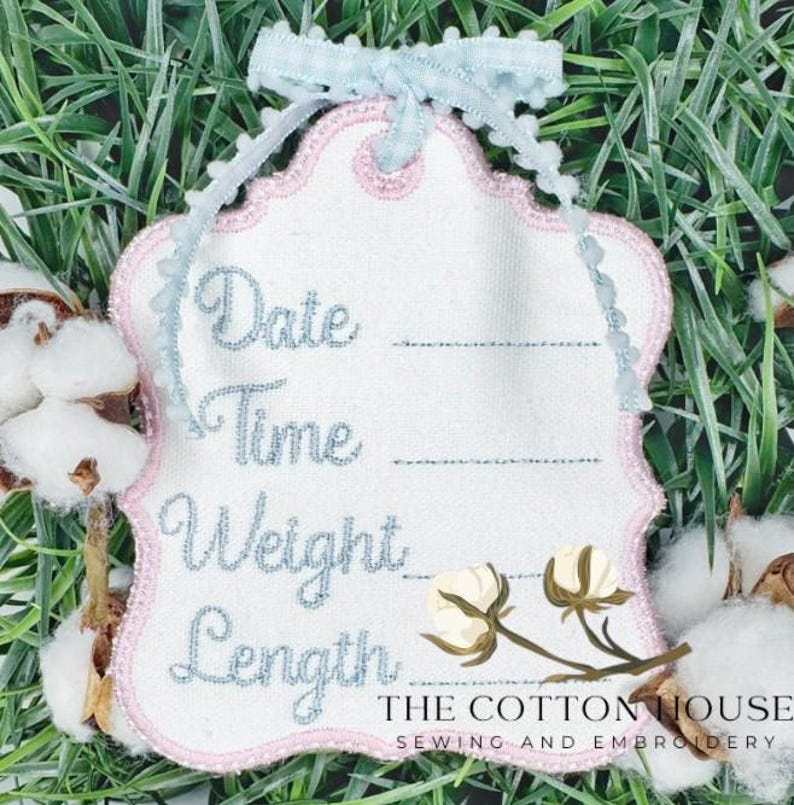 May include: White embroidered tag with pink trim and light blue text reading "Date, Time, Weight, Length." A light blue ribbon and cotton sprigs accent the tag. The text "THE COTTON HOUSE SEWING AND EMBROIDERY" is at the bottom.