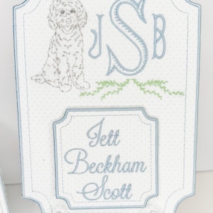 May include: Embroidered white fabric plaque with a light blue border and polka dot pattern. Features a dog illustration, initials, and the name "Jett Beckham Scott" in script. Includes a clear plastic stand.